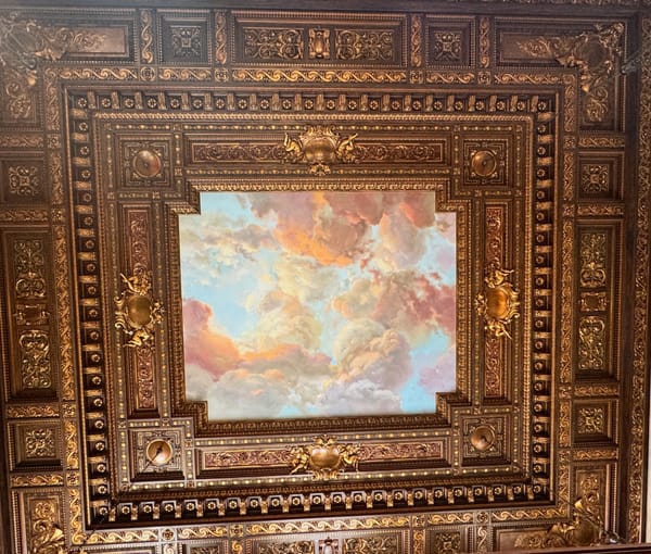 A photo of a beautiful ceiling with ornaments and a painting of clouds.